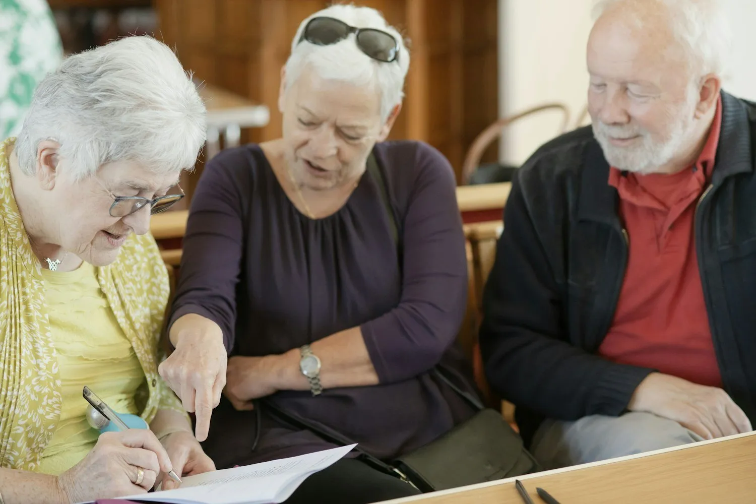 Three older adults leaning over a document together, one pointing at it.