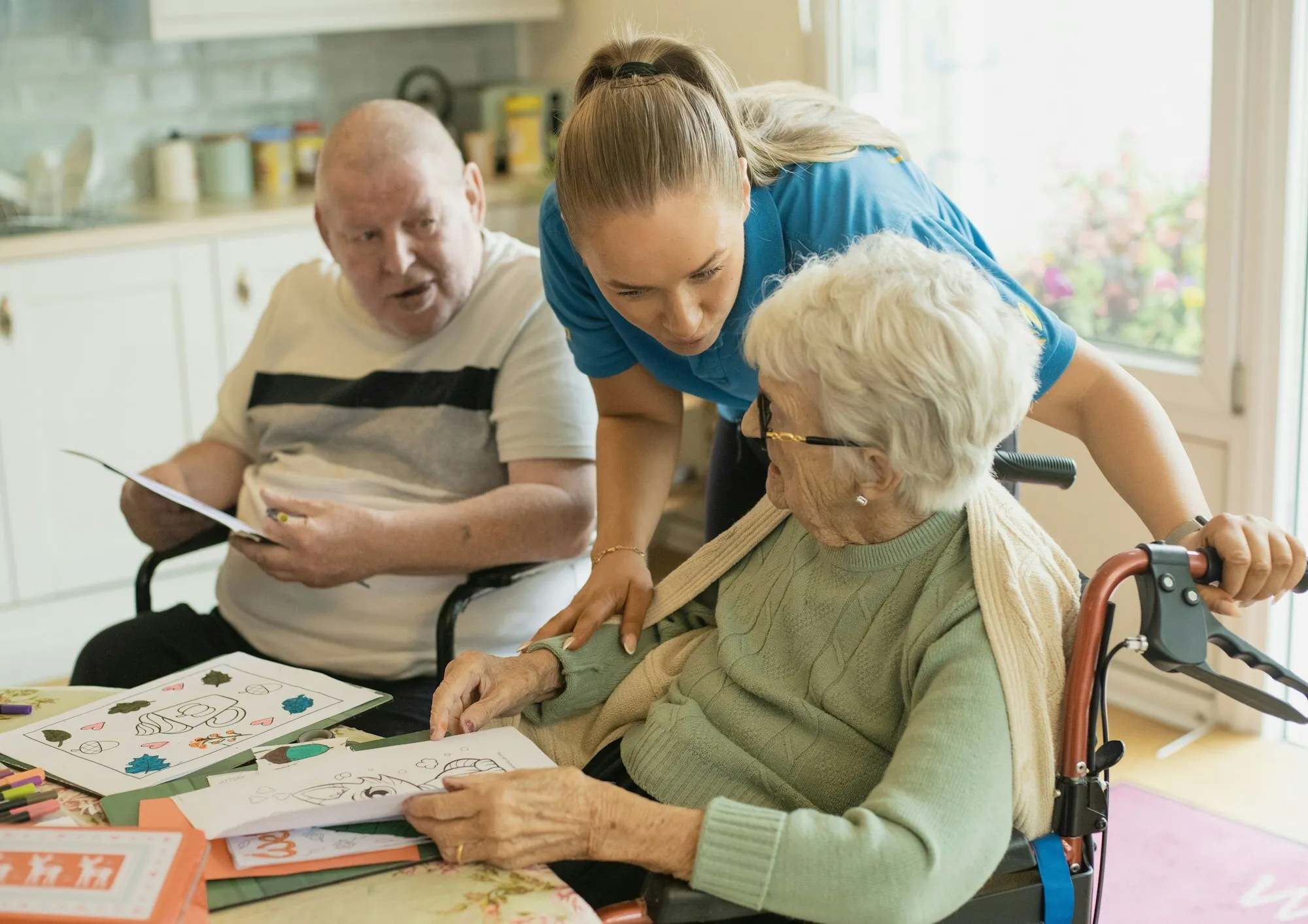 A caregiver helping an older couple at their kitchen table, a walker and wheelchair beside them.