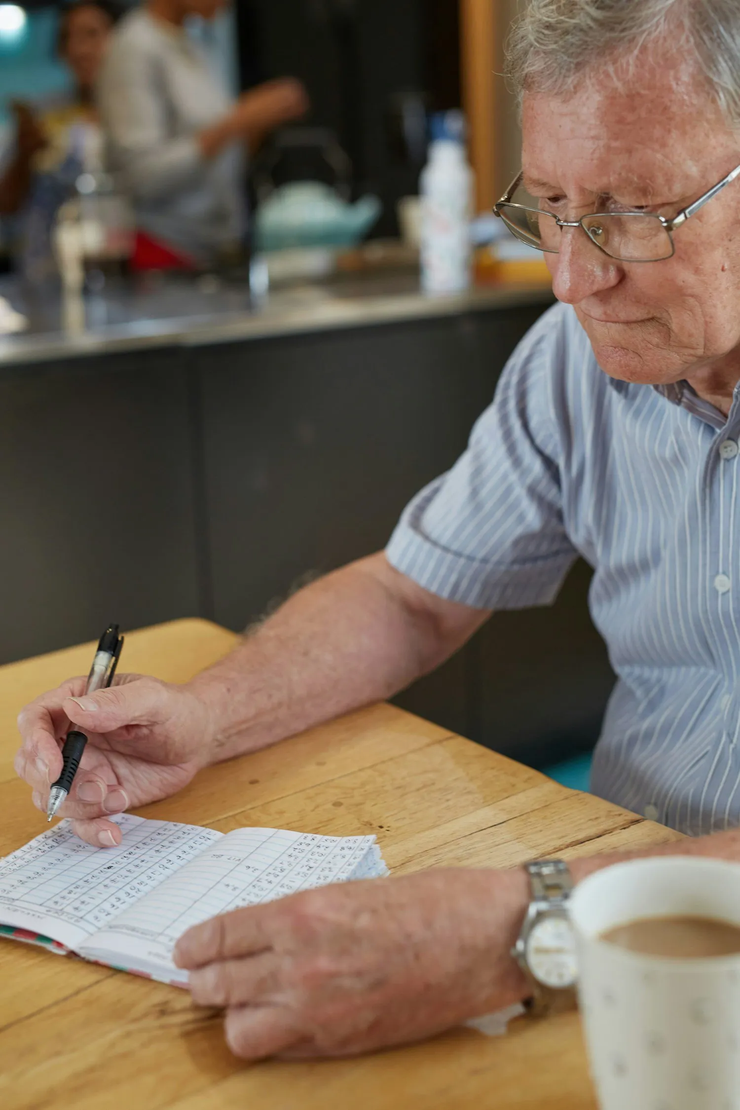 An older man in glasses filling out a form at a table, a cup of coffee beside him.